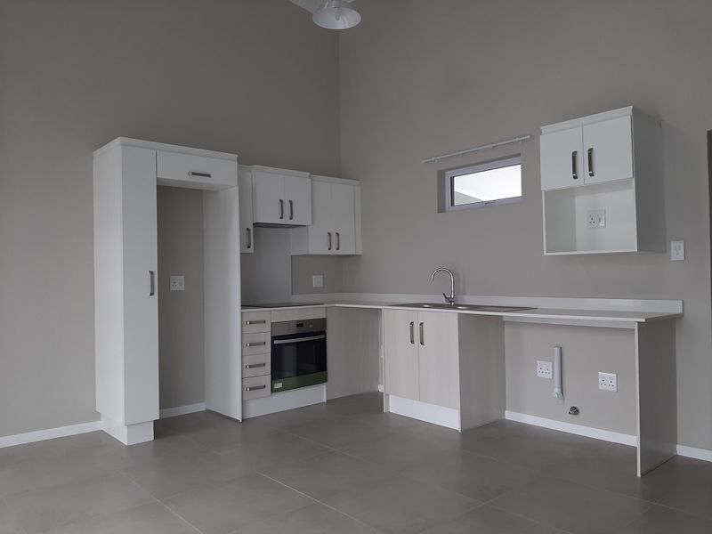 A clean, modern, and empty kitchen featuring white cabinetry, integrated oven, hob, and stainless steel sink with tiled flooring.