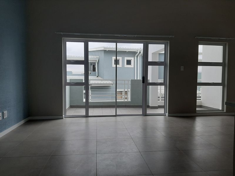 An empty, well-lit living area featuring large sliding glass doors that open onto a balcony, with neutral tiled flooring throughout.