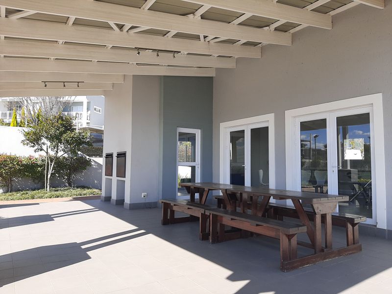A spacious covered patio featuring tiled flooring, exposed beam ceiling, and built-in braai facilities, providing seamless access to the interior through double glass doors.
