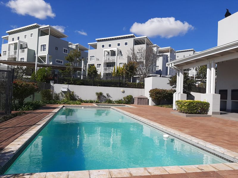 A well-maintained communal swimming pool area featuring paved decking, a covered patio space, and landscaped surroundings within a modern apartment complex.