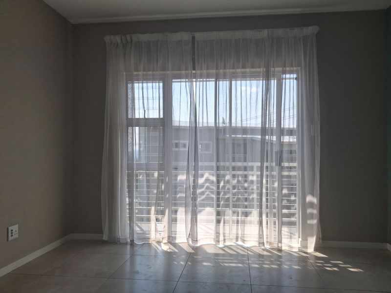 An empty living room featuring neutral tiled flooring and a large sliding glass door with sheer curtains, providing access to an outdoor balcony area.
