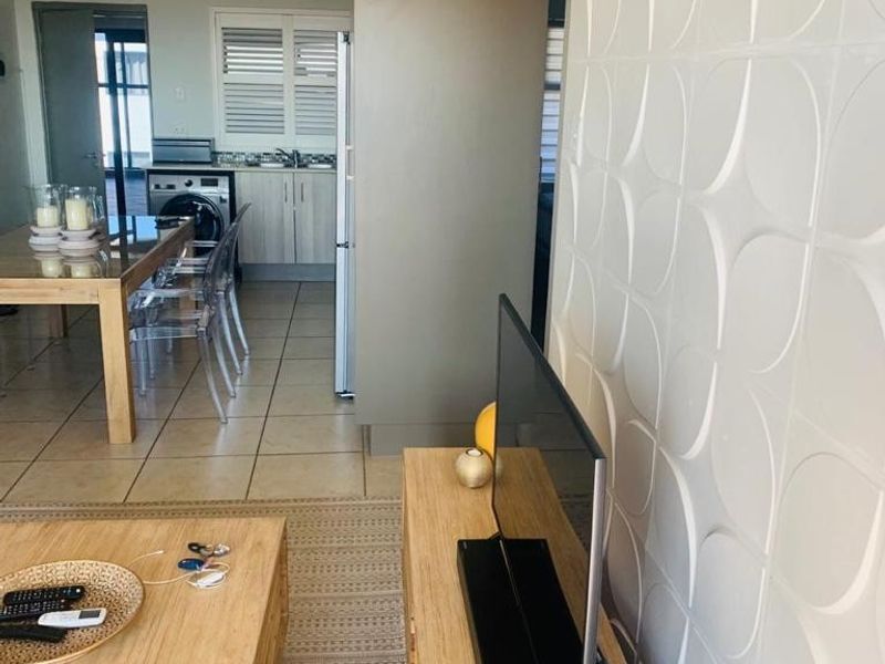 Open plan living area featuring a textured feature wall, ceramic floor tiles, and a functional kitchen space with cabinetry and shutter blinds.