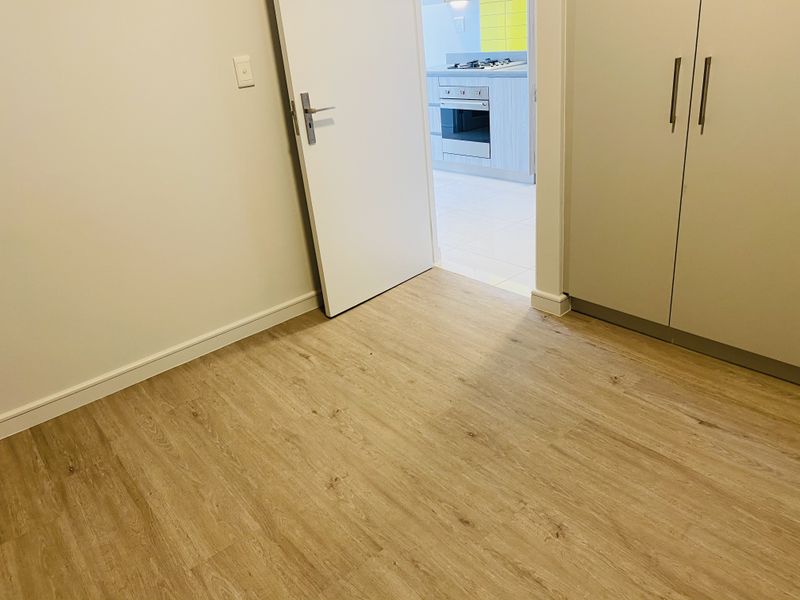 An empty bedroom featuring light wood-look laminate flooring and built-in cupboards, with a view through the doorway into the adjacent kitchen area.