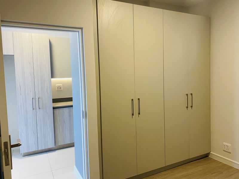 A clean, modern bedroom featuring ample built-in cupboard storage, light wood-look laminate flooring, and a view into an adjacent tiled kitchen area.