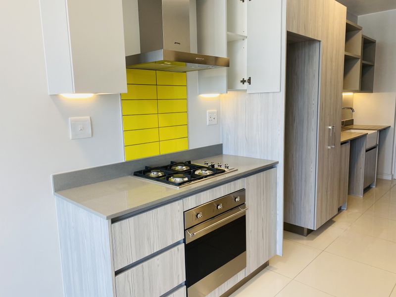Modern kitchen featuring light wood-grain cabinetry, stone countertops, a gas hob with extractor fan, and a vibrant yellow tiled backsplash.
