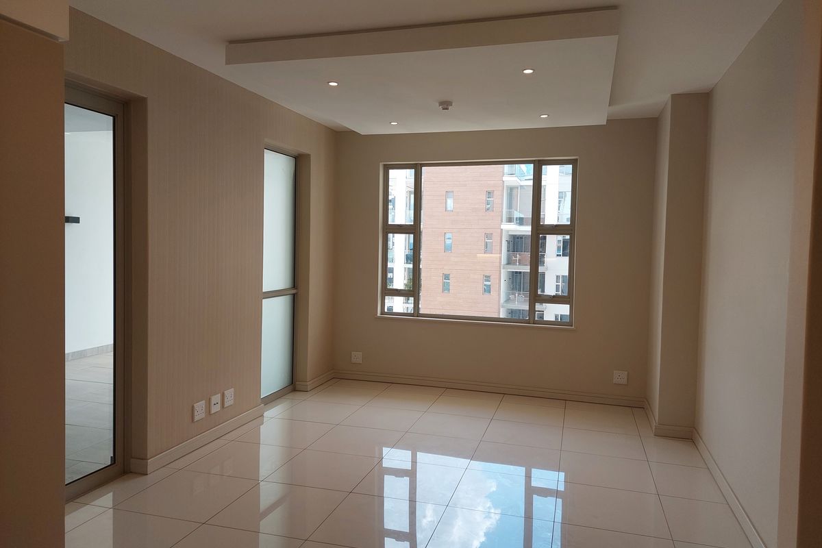 A bright, empty living area featuring modern tiled flooring, a recessed ceiling with downlights, and large windows providing natural light.