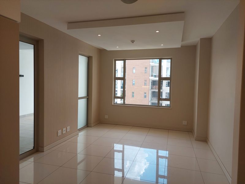 A bright, empty living area featuring modern tiled flooring, a recessed ceiling with downlights, and large windows providing natural light.