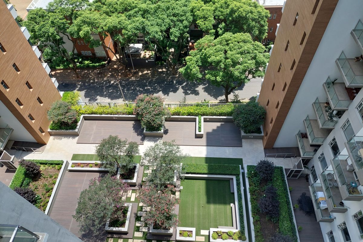 An elevated aerial view of a well-maintained apartment complex courtyard featuring landscaped gardens, timber decking, artificial grass, and paved pathways.