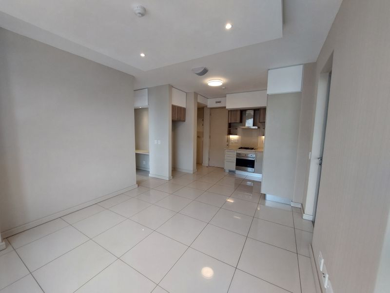 A bright, open-plan living and kitchen area featuring neutral tiled flooring, modern recessed lighting, and a compact, well-maintained kitchen with built-in appliances.