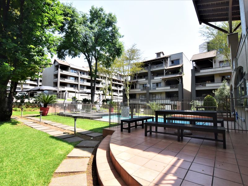 Communal outdoor area featuring a swimming pool, tiled patio space, and landscaped garden within a well-maintained apartment complex.