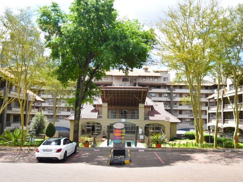 Front view of a multi-story apartment complex featuring a paved driveway, mature trees, and balcony access for residents.