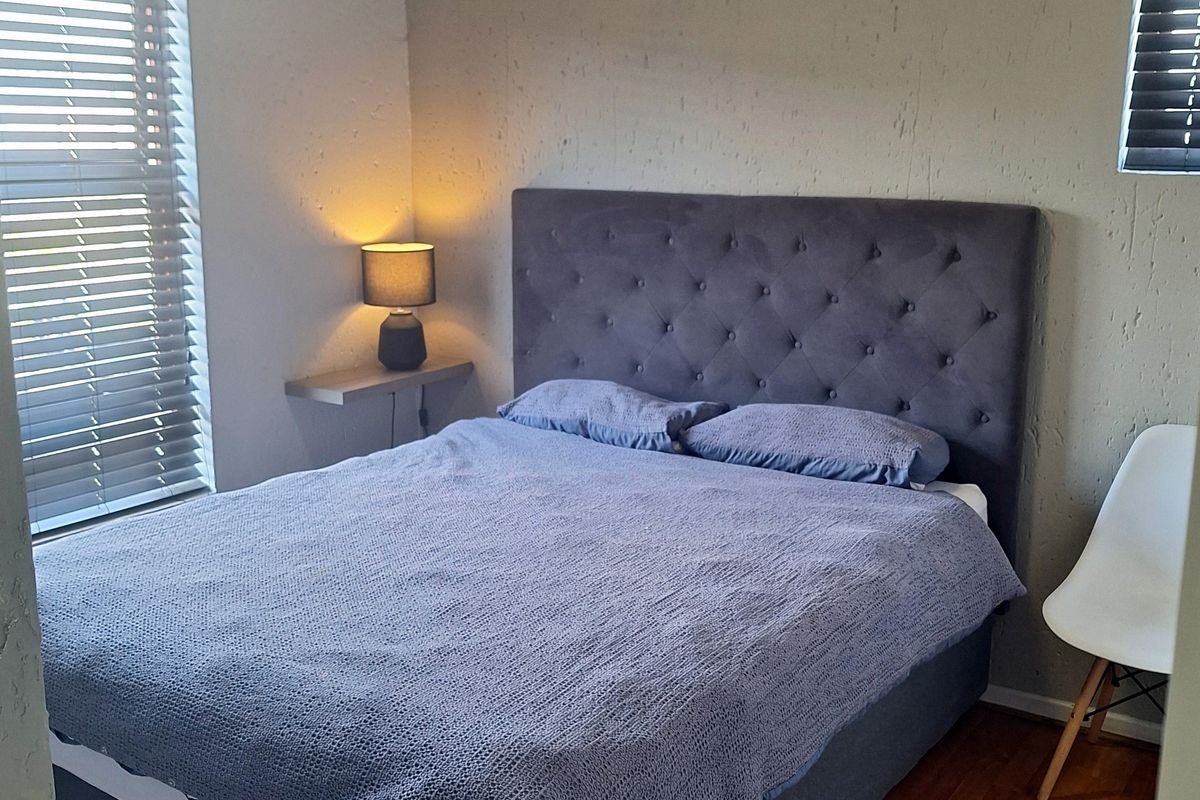 A functional bedroom featuring laminate flooring, dual windows with venetian blinds, and a built-in floating bedside shelf.