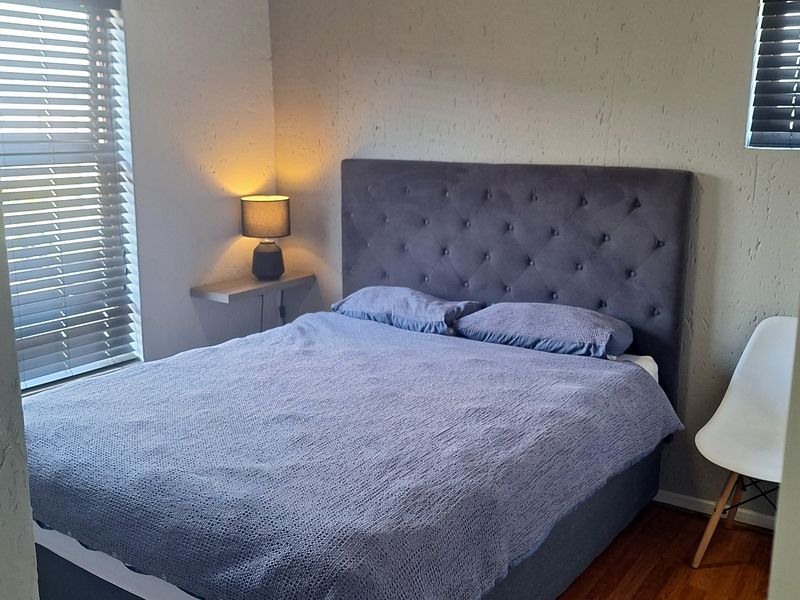 A functional bedroom featuring laminate flooring, dual windows with venetian blinds, and a built-in floating bedside shelf.
