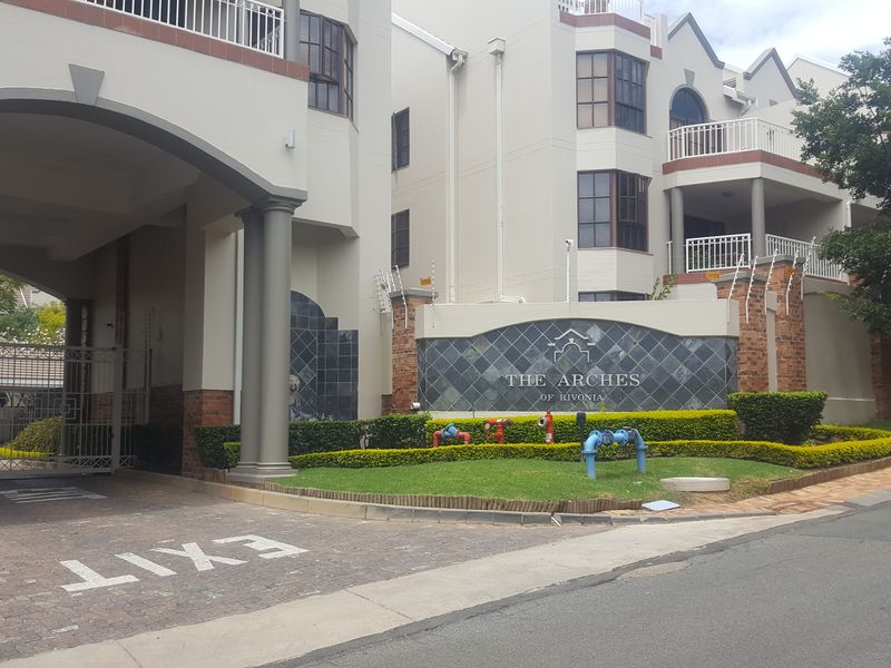Front entrance view of The Arches of Rivonia apartment complex, featuring a secure gated access, brick paving, and well-maintained landscaped gardens.