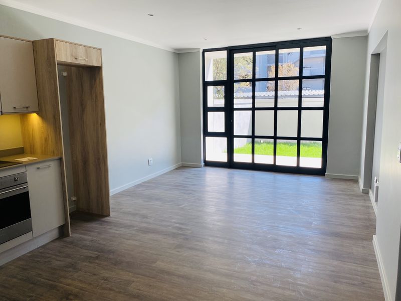 Modern open-plan living area featuring light wood-tone cabinetry, integrated oven, laminate flooring, and large black-framed glass doors opening to a private garden space.