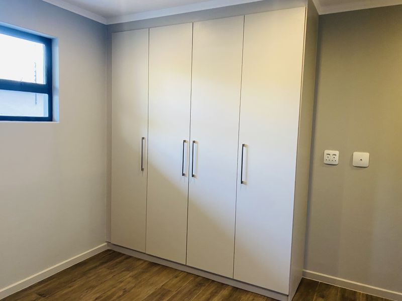A clean, modern bedroom featuring neutral walls, wood-look laminate flooring, and a large, white built-in wardrobe with sleek handles.