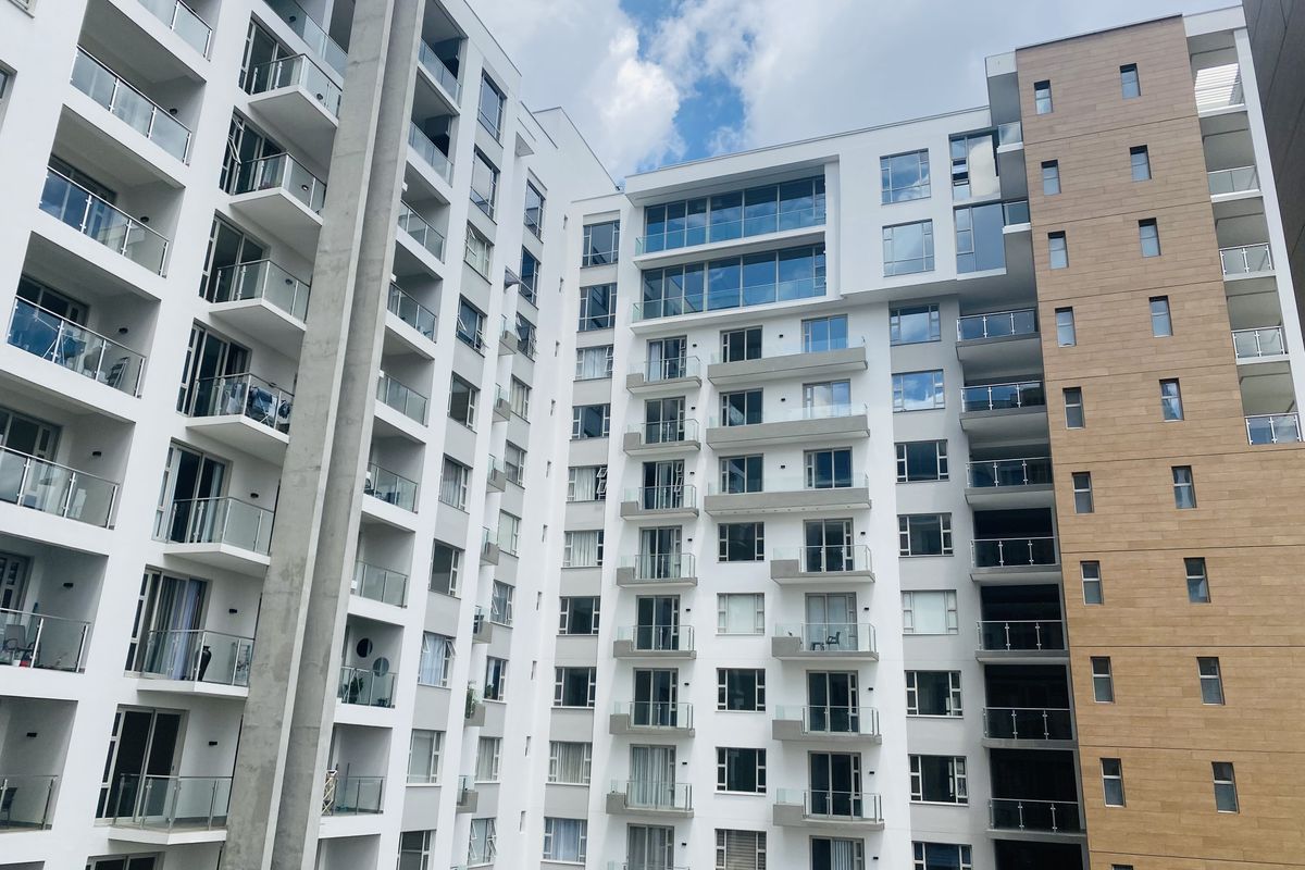 A modern apartment building exterior featuring multiple levels of balconies with glass balustrades and a mix of white concrete and wood-look cladding.
