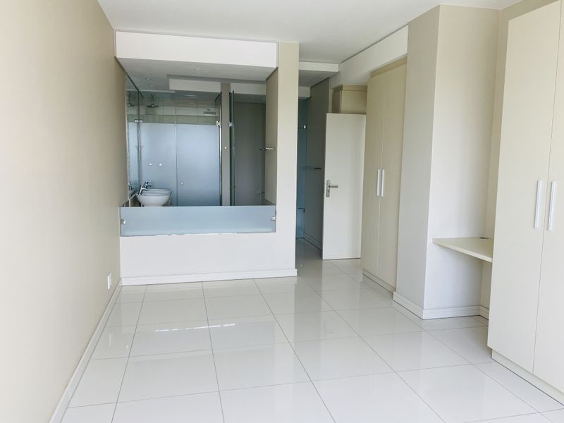 Spacious bedroom featuring tiled flooring, extensive built-in cupboards, a built-in desk, and an open-plan en-suite bathroom with a glass shower partition.