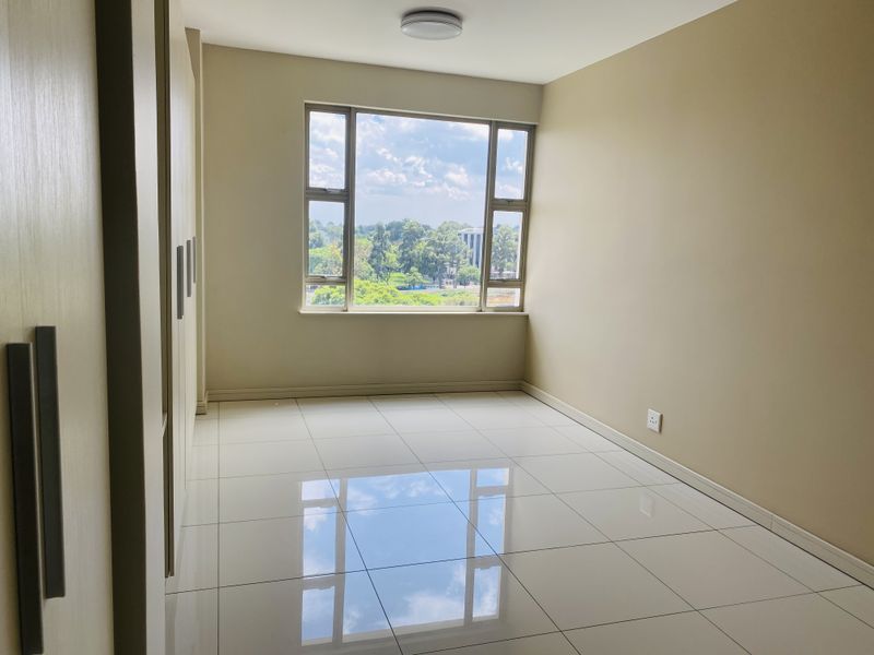 A bright, empty bedroom featuring polished tiled flooring, built-in cupboards, and a large window offering pleasant outdoor views.
