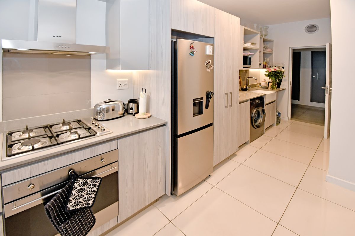 Modern kitchen featuring light wood-grain cabinetry, stone countertops, gas hob, built-in oven, extractor fan, and a farmhouse sink with integrated laundry area.