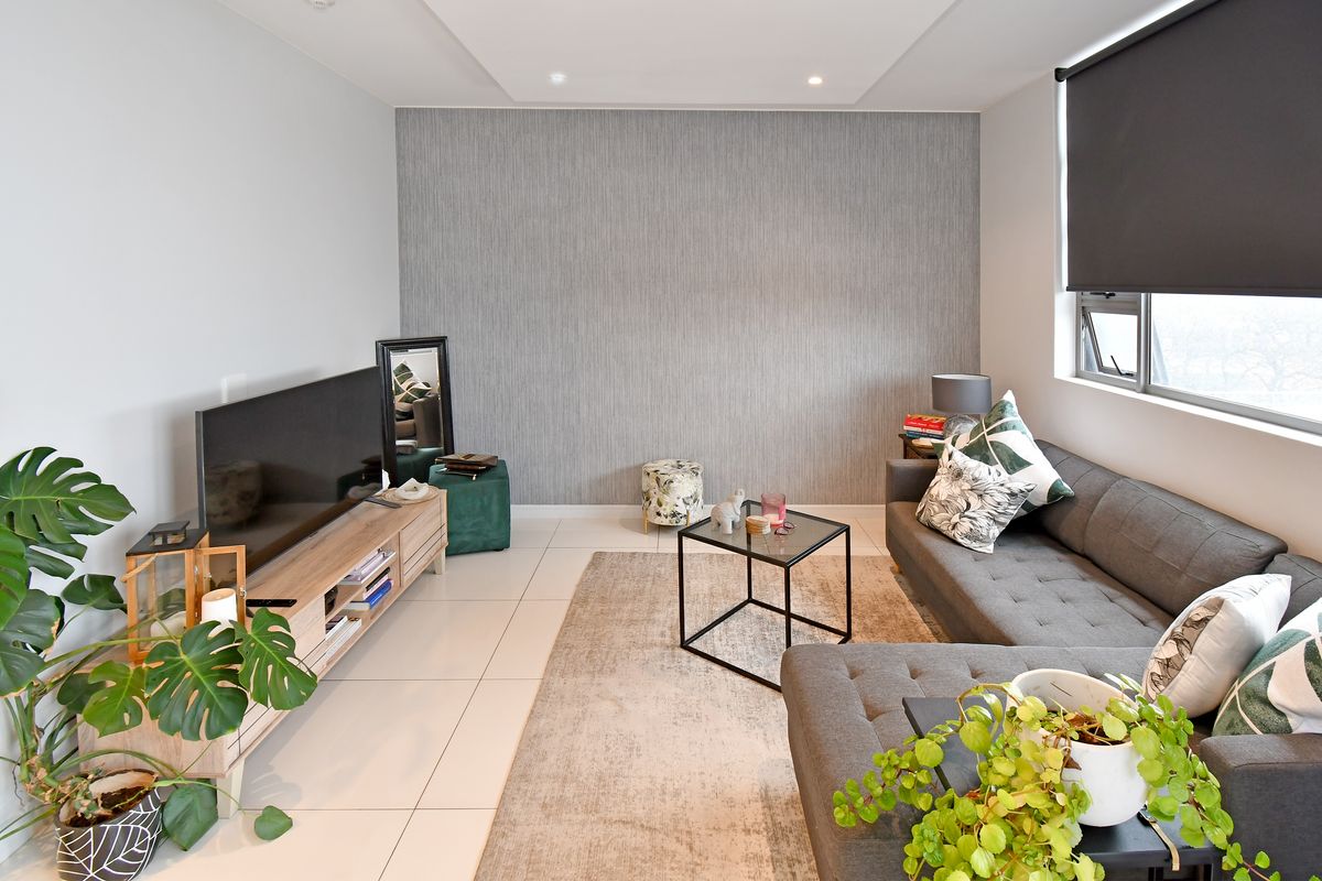 A well-maintained living room featuring neutral tiled flooring, recessed downlighting, a textured feature wall, and modern roller blinds.