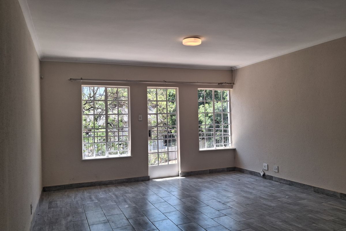 A bright, empty living room featuring modern grey tiled flooring, large windows, and a central glass-paneled door providing access to the exterior.