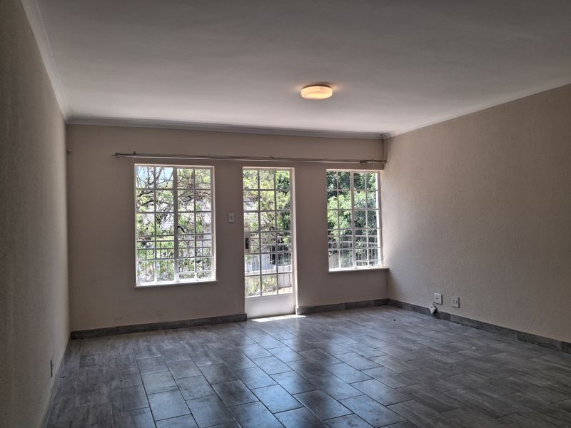 A bright, empty living room featuring modern grey tiled flooring, large windows, and a central glass-paneled door providing access to the exterior.