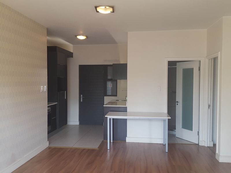 An open-plan living area featuring laminate flooring, a compact kitchen with dark cabinetry, and access to a bathroom through a frosted glass door.