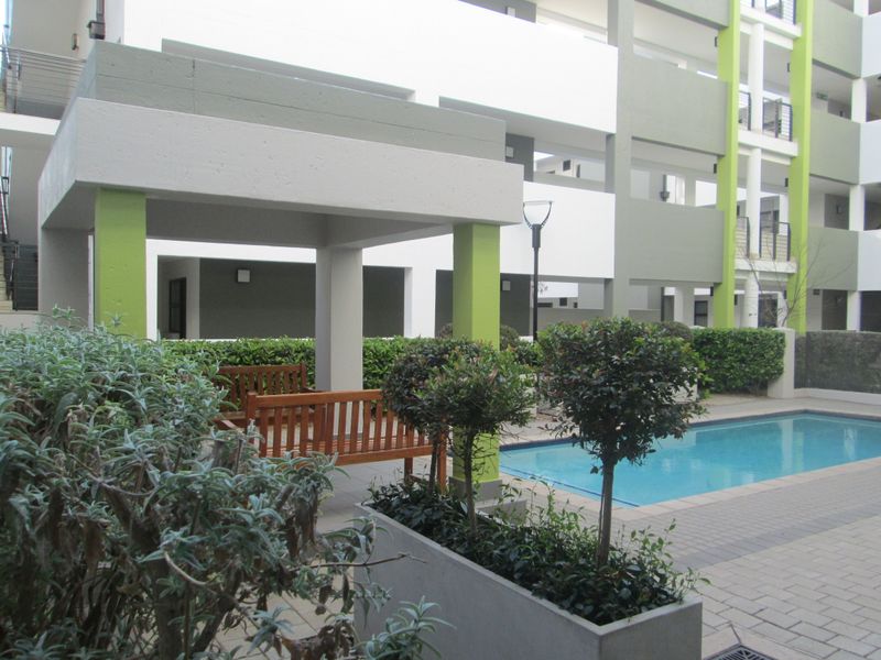 A well-maintained communal pool area featuring a paved patio, a covered pergola, and established garden planters within an apartment complex.