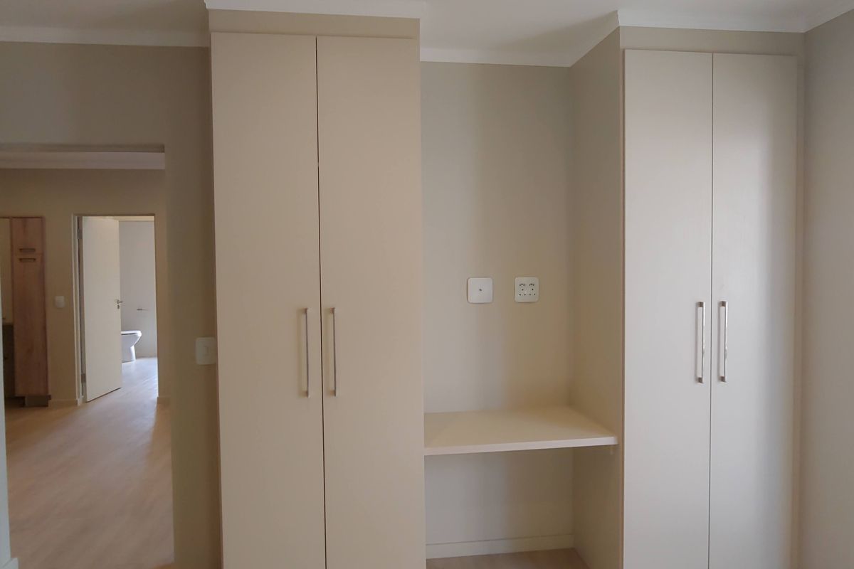 A bright, modern bedroom featuring neutral-toned built-in cupboards with an integrated desk area, laminate flooring, and clean finishes throughout.