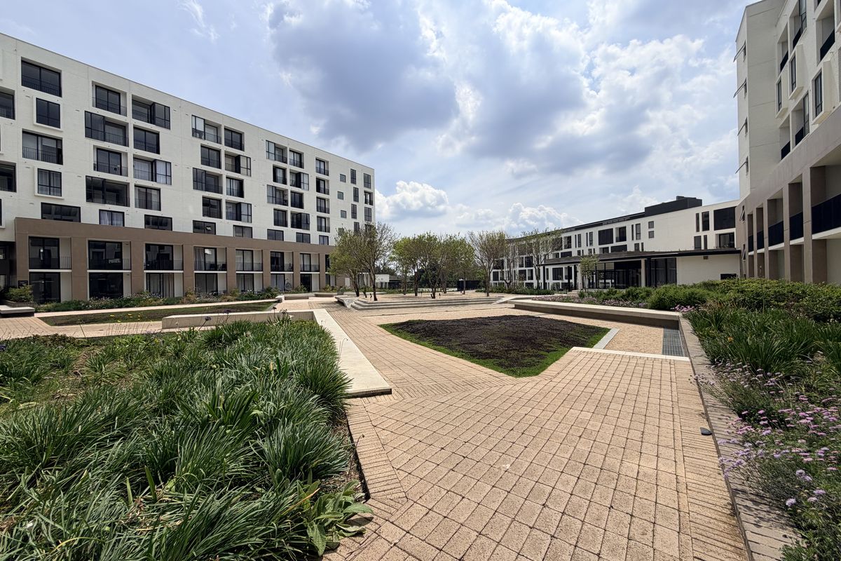 A well-maintained communal courtyard area featuring paved walkways, landscaped garden beds, and modern apartment building facades.