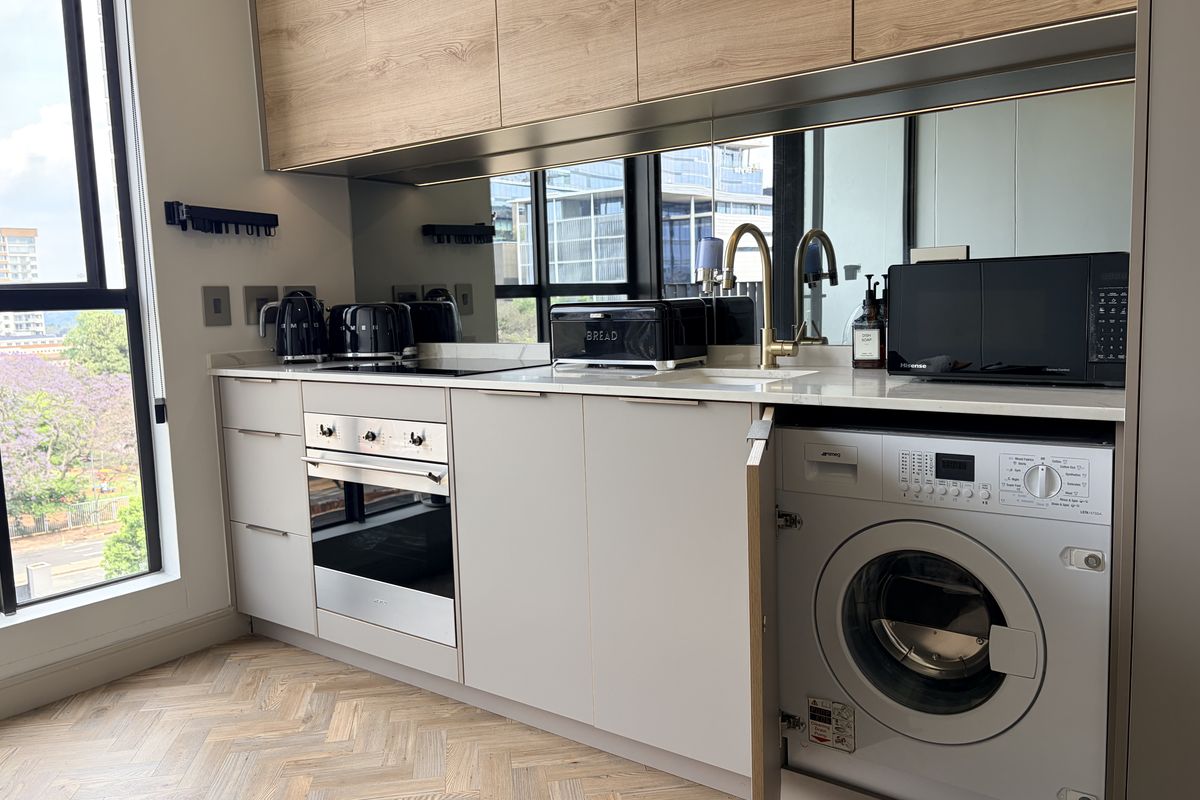 Modern kitchen featuring light cabinetry, stone countertops, mirrored splashback, integrated appliances, and elegant herringbone timber flooring with large windows providing natural light.