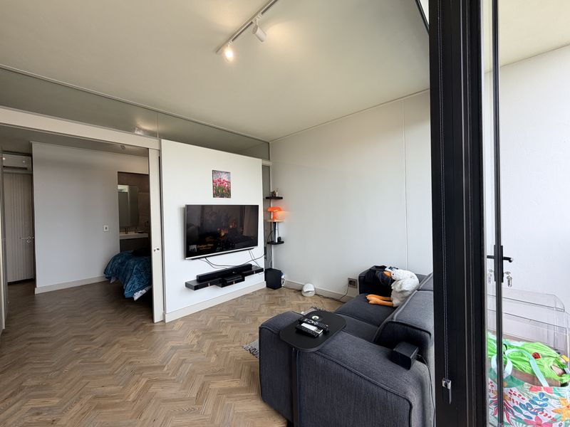 A modern living room featuring herringbone timber flooring, track lighting, and a sliding glass door leading to an outdoor area, with an adjacent bedroom view.