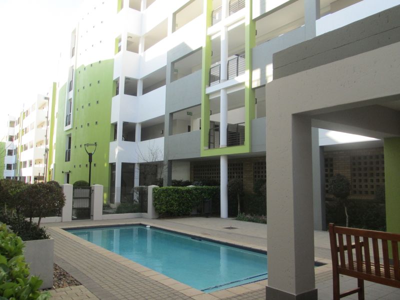 A well-maintained communal swimming pool area set within a modern multi-story apartment complex with paved decking and landscaped garden features.