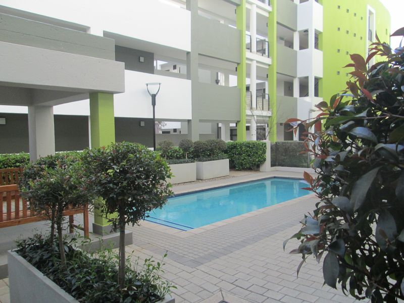 A well-maintained communal swimming pool area featuring paved decking and integrated landscaping planters within a modern apartment complex.