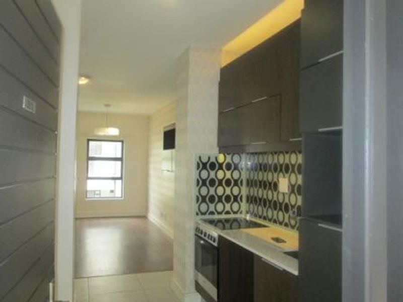 An open-plan apartment layout featuring a modern kitchen with dark cabinetry, built-in appliances, and a patterned backsplash, leading into a bright living area.