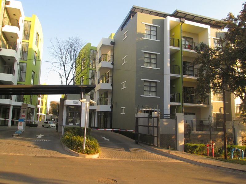 Modern apartment complex entrance featuring a secure gated access point, guard house, paved driveway, and multi-story residential buildings with private balconies.