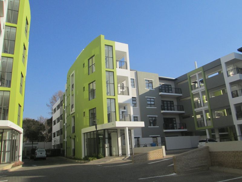 A modern apartment complex featuring a vibrant green and grey facade with multiple balconies and a paved driveway entrance.