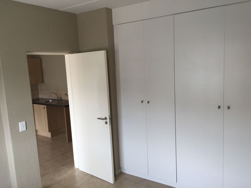 A clean, empty bedroom featuring neutral wall tones, tiled flooring, and ample built-in cupboard storage, with a view into the adjacent kitchen area.