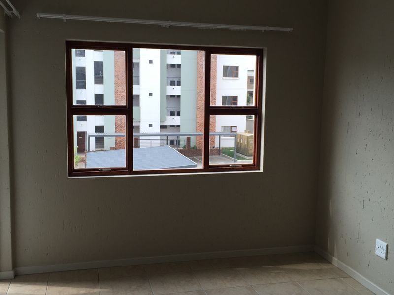 An empty bedroom featuring tiled flooring, a ceiling-mounted spotlight fixture, and a window with a brown frame overlooking the apartment complex.