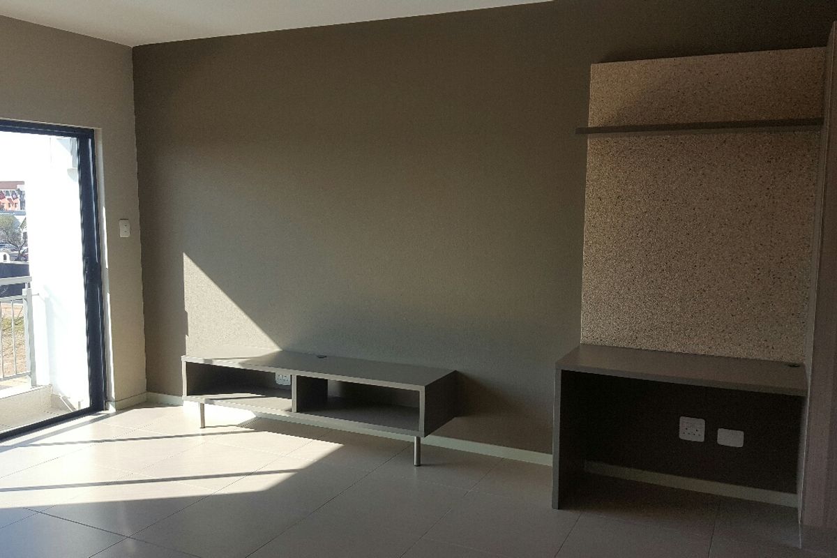 A bright, empty living area featuring neutral tiled floors, a built-in media console, a dedicated desk space, and access to a balcony.