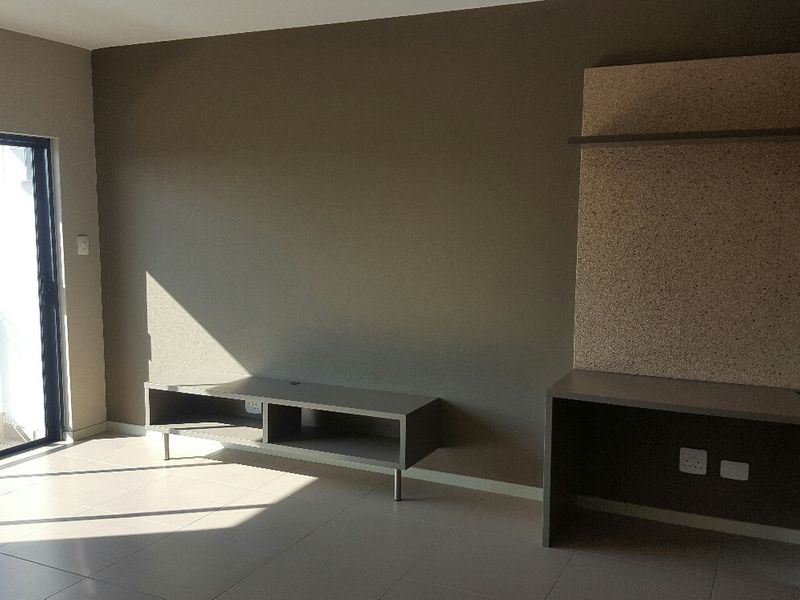 A bright, empty living area featuring neutral tiled floors, a built-in media console, a dedicated desk space, and access to a balcony.