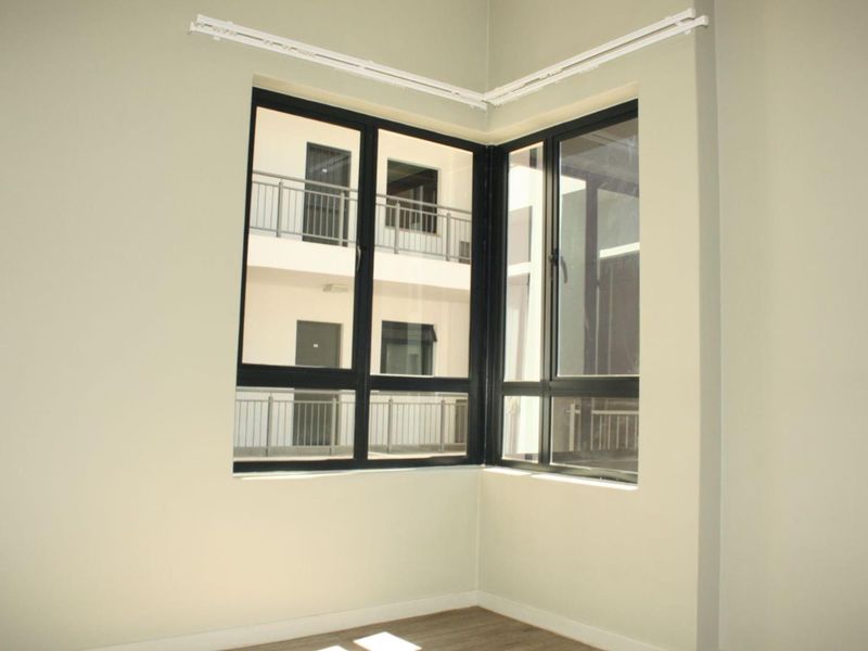 A bright, empty bedroom featuring a modern corner window with black aluminum frames and a neutral wall finish.