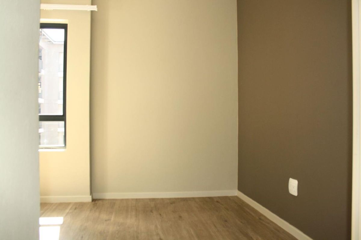 A bright, empty bedroom featuring modern laminate flooring, neutral wall tones, and a large window providing natural light.
