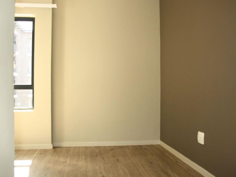 A bright, empty bedroom featuring modern laminate flooring, neutral wall tones, and a large window providing natural light.