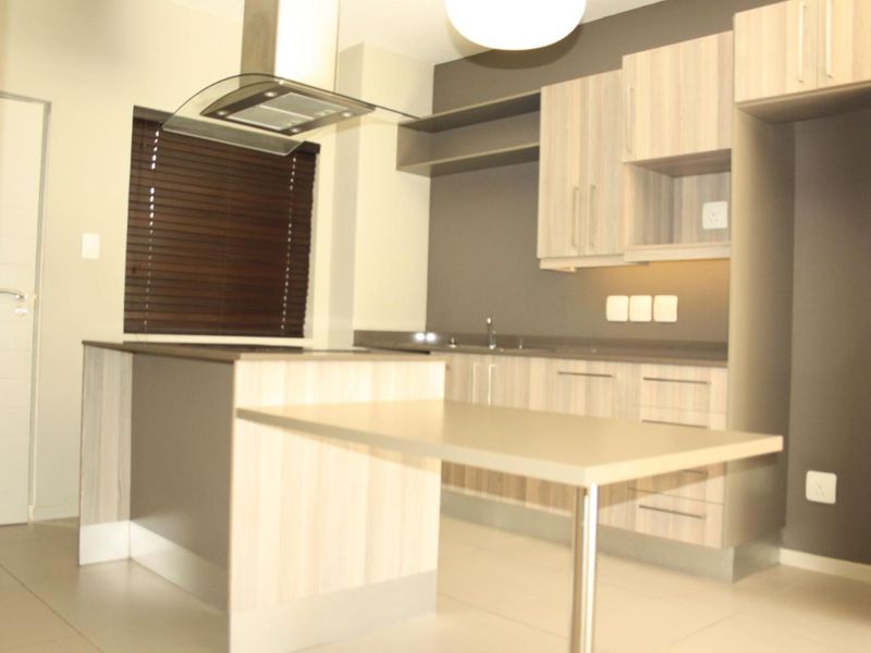 Modern kitchen featuring light wood-finish cabinetry, a central island with breakfast bar, integrated extractor fan, and neutral tiled flooring.
