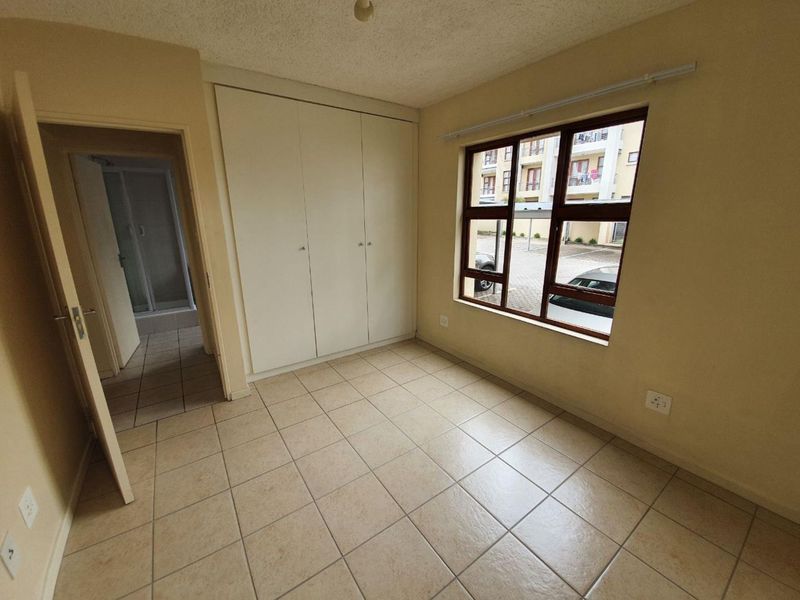 An empty bedroom featuring tiled flooring, built-in cupboards, and an aluminium window frame, with an adjacent bathroom visible through the doorway.