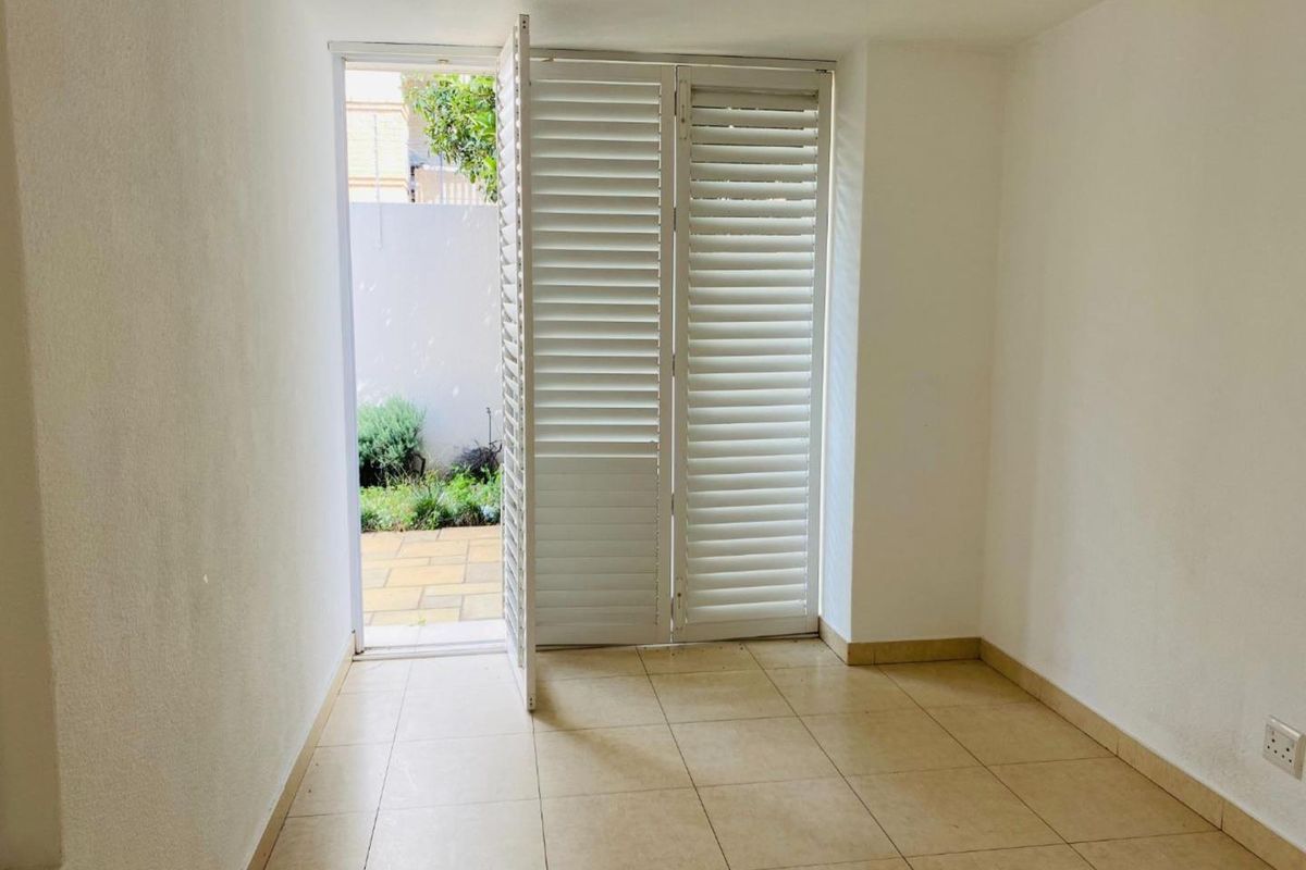 An empty, well-lit living area featuring neutral tiled flooring and white plantation shutters providing direct access to an outdoor patio space.