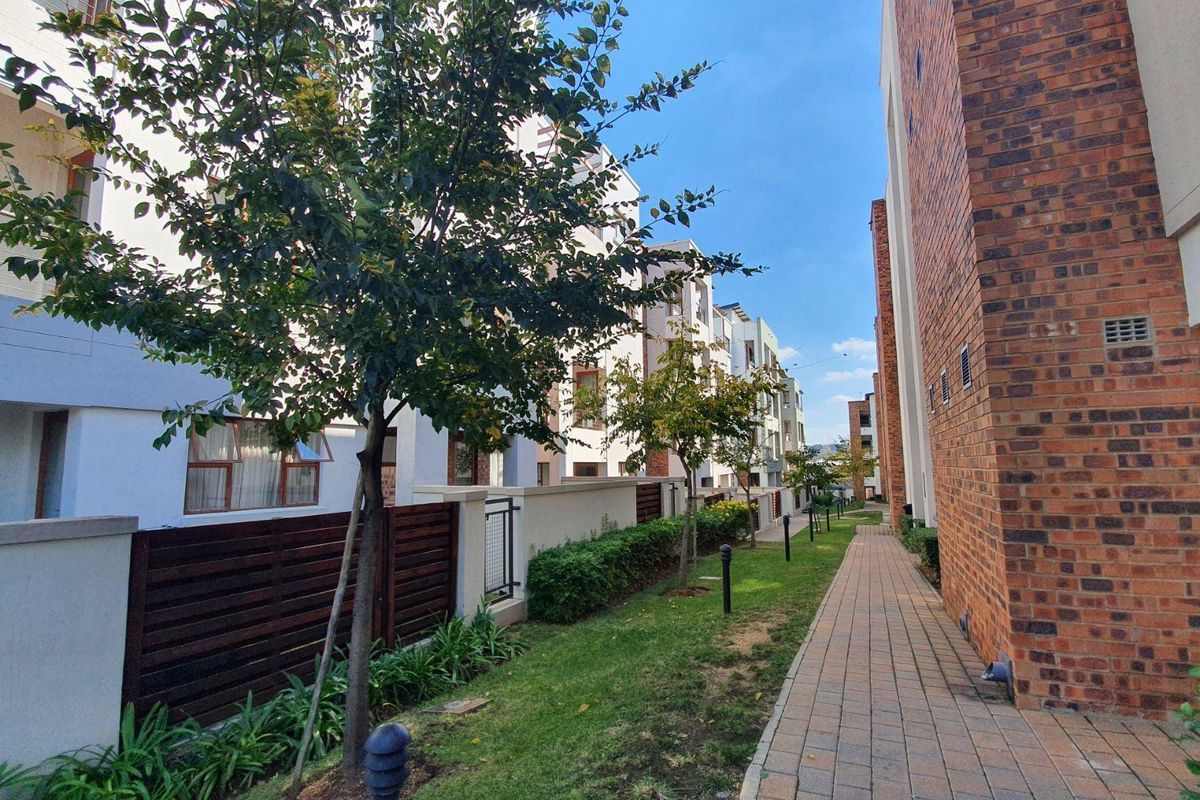 A well-maintained exterior walkway between apartment blocks, featuring brick facades, paved paths, and landscaped garden areas with integrated lighting.
