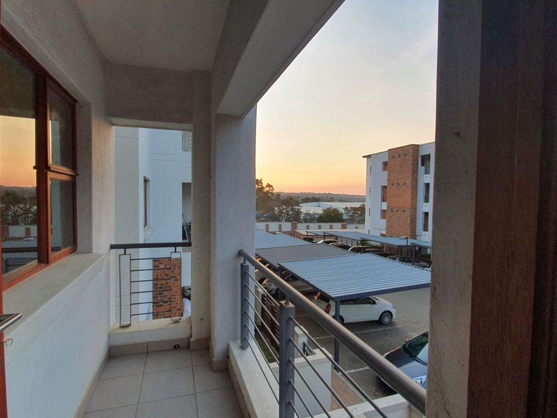A covered balcony featuring tiled flooring and a metal balustrade, offering an elevated view of the apartment complex parking area.